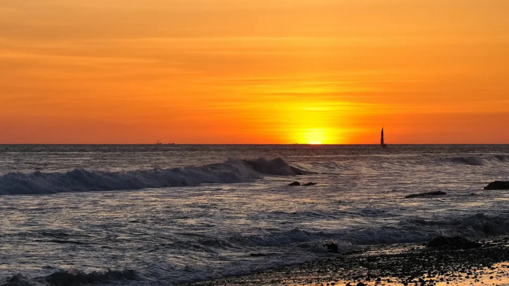 Ocean waves at sunset with a sailboat on the horizon.