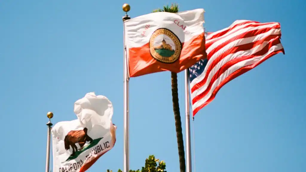 California and U.S. flags waving on flagpoles against a blue sky.